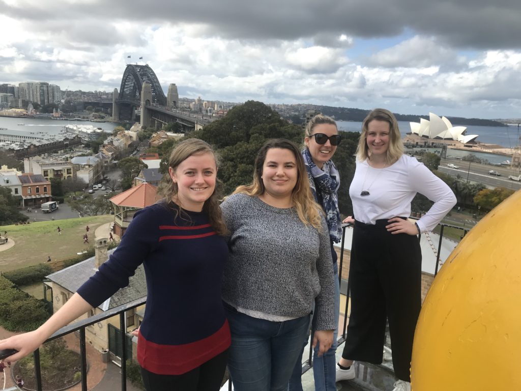 Four women pose for a photo on a high balcony with the Sydney Harbour Bridge and the Opera House in the background. In the foreground is visible part of a very large yellow ball.